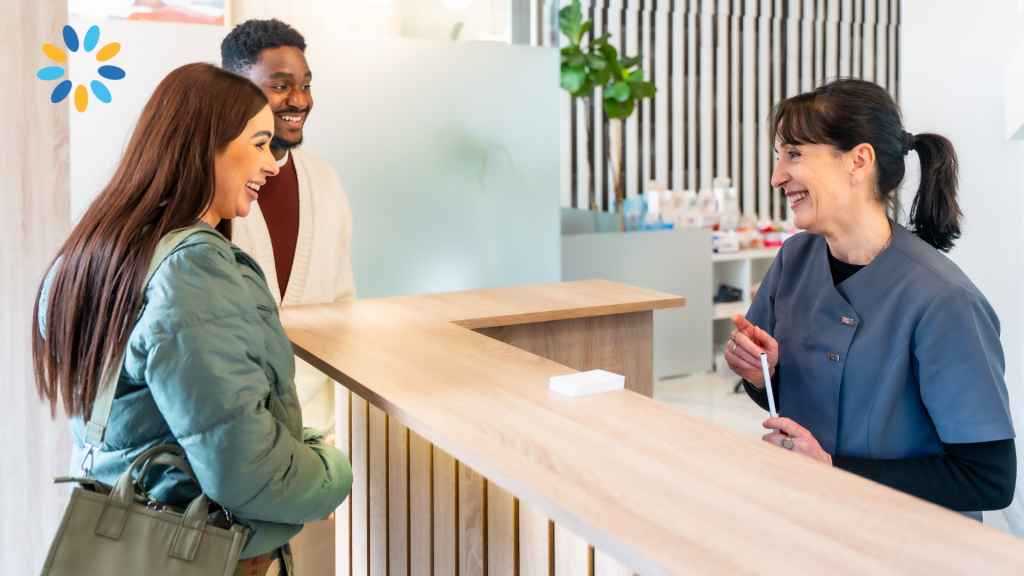 A member of reception staff smiles while speaking with two patients at a modern general practice reception desk. The patients are smiling and engaged in conversation, illustrating a welcoming and positive patient experience. The Primary Care Training Hub logo appears in the top left corner.